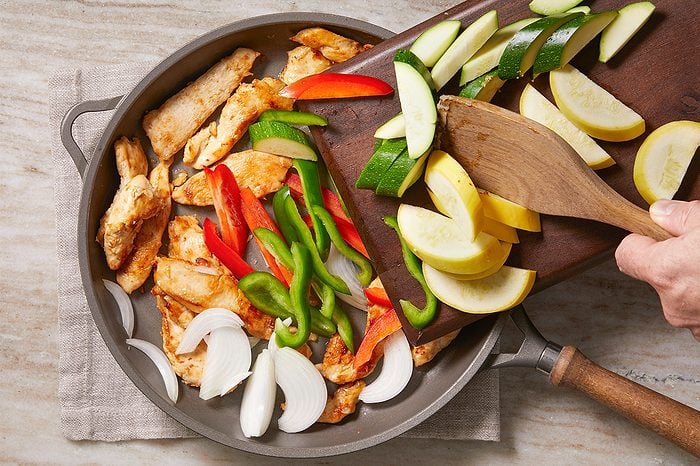 Overhead horizontal image of a hand sliding sliced yellow squash, zucchini, red pepper, and green pepper from a wooden cutting board into a skillet. The skillet contains cooked chicken strips and thinly sliced onions, preparing for the next stage of cooking.