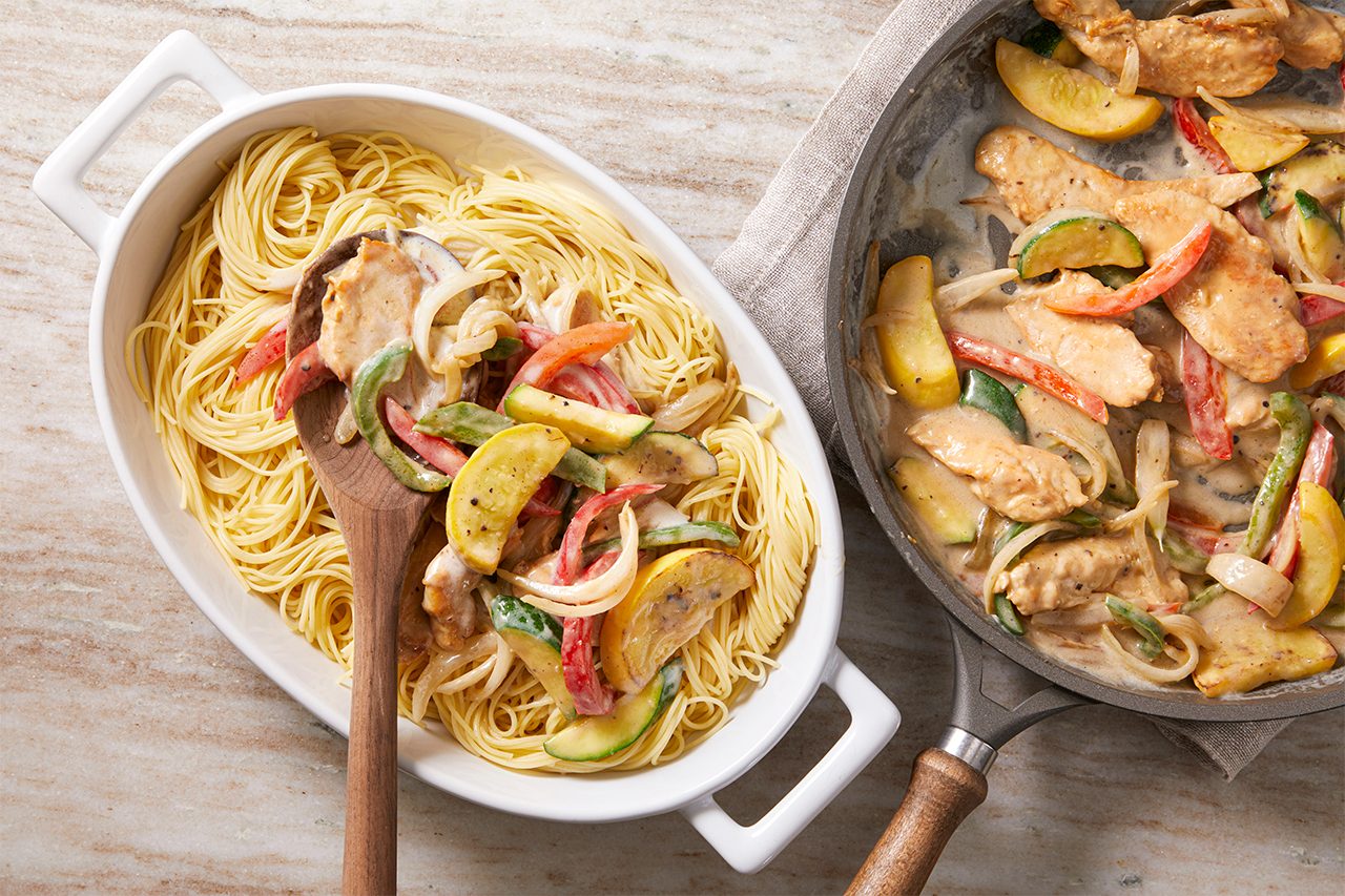 Overhead horizontal image of cooked angel hair pasta in a white serving dish, being mixed with a wooden spoon into a creamy sauce containing chicken strips, yellow squash, zucchini, red bell pepper, green bell pepper, and onions. A skillet to the right holds more of the chicken and vegetable mixture in the sauce.