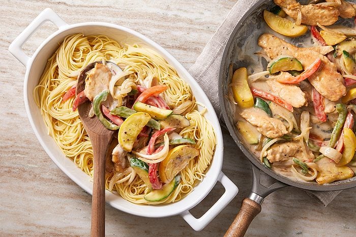 Overhead horizontal image of cooked angel hair pasta in a white serving dish, being mixed with a wooden spoon into a creamy sauce containing chicken strips, yellow squash, zucchini, red bell pepper, green bell pepper, and onions. A skillet to the right holds more of the chicken and vegetable mixture in the sauce.