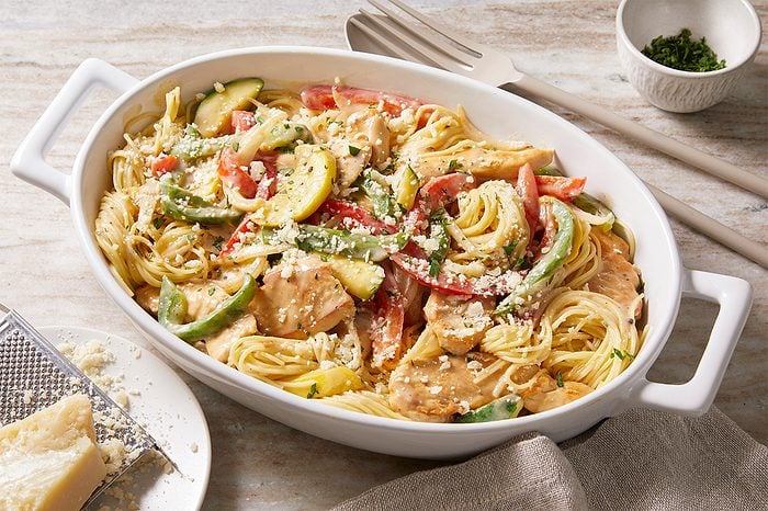 Eye Level horizontal image of Chicken Garden Medley, a pasta dish with chicken and vegetables, served in a white oval baking dish. The dish is topped with shredded Parmesan cheese and fresh herbs, with a block of Parmesan and a grater visible nearby.