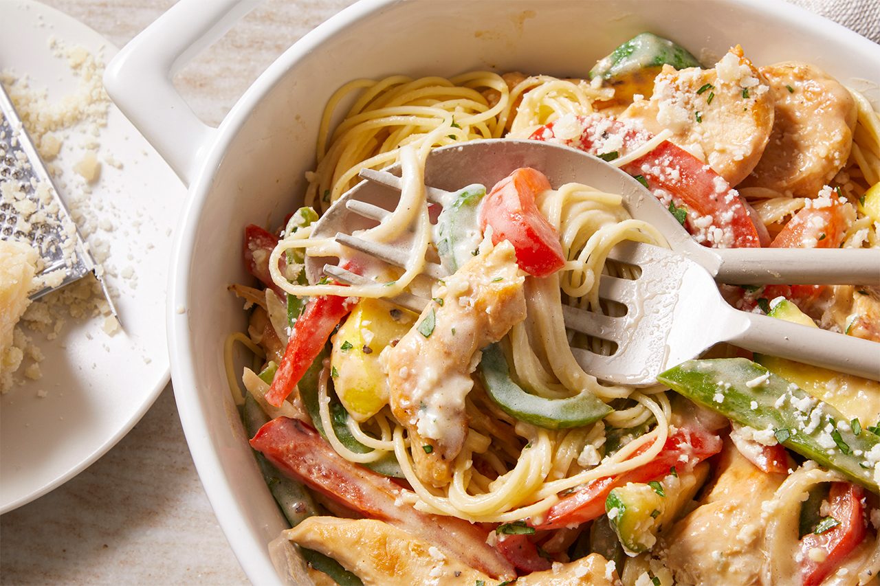 Overhead horizontal image of a white serving dish filled with Chicken Garden Medley. Cooked angel hair pasta, chicken strips, sliced yellow squash, zucchini, and julienned red and green peppers are coated in a creamy sauce and topped with shredded Parmesan cheese. A serving utensil rests in the dish, with a block of Parmesan cheese and a grater visible beside it.
