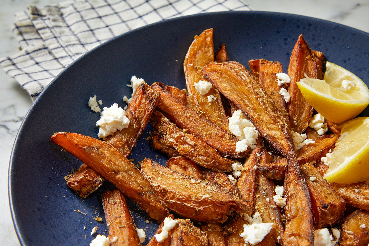 Eye Level horizontal image of roasted potato wedges seasoned with herbs and crumbled feta cheese, served on a dark blue plate. Two fresh lemon wedges accompany the dish, with a checkered napkin visible in the background.