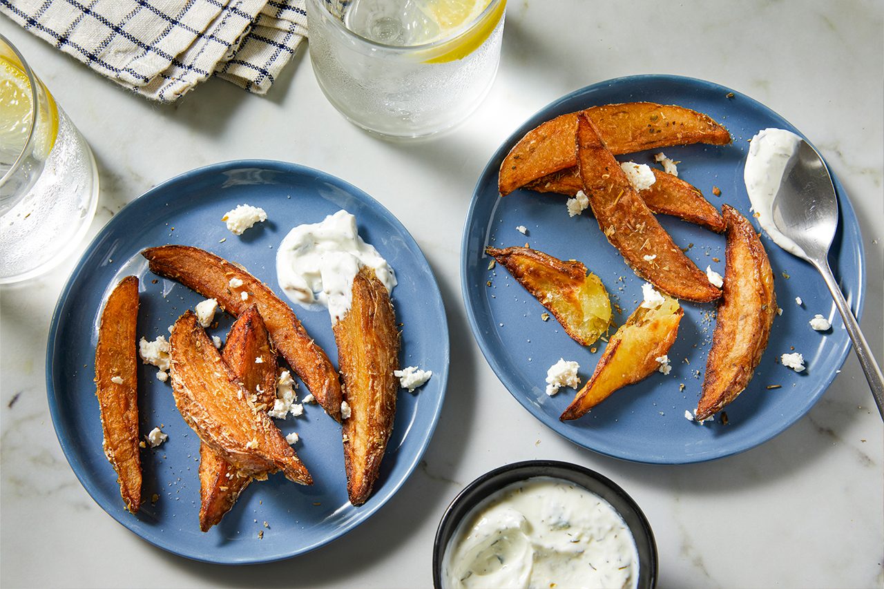 Overhead horizontal image of two blue plates filled with roasted potato wedges. The wedges are seasoned and topped with crumbled feta cheese, served alongside a creamy dipping sauce. Two glasses of lemon water and an additional bowl of dipping sauce are also visible on a marble surface.