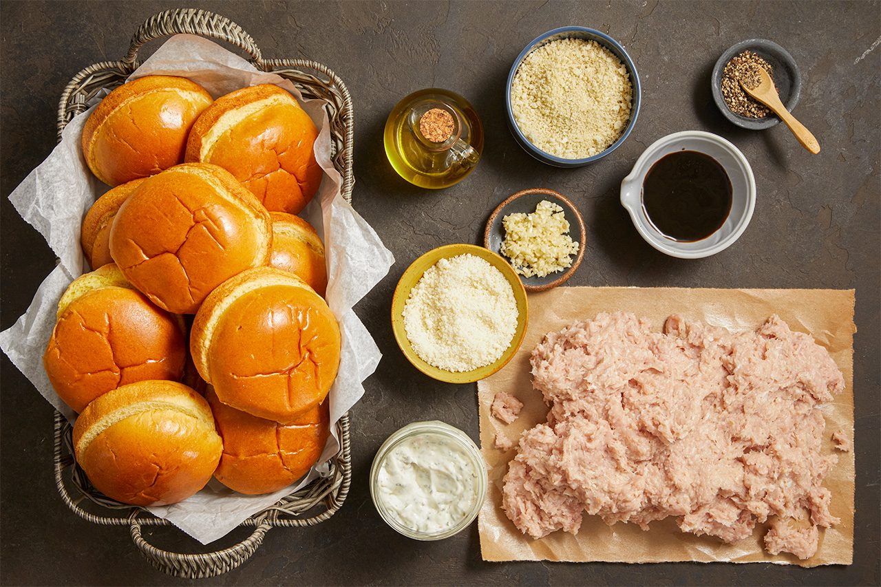Top-down view of a basket of hamburger buns, a pile of raw ground meat, and small bowls containing oil, breadcrumbs, herbs, shredded cheese, and seasonings on a dark surface.