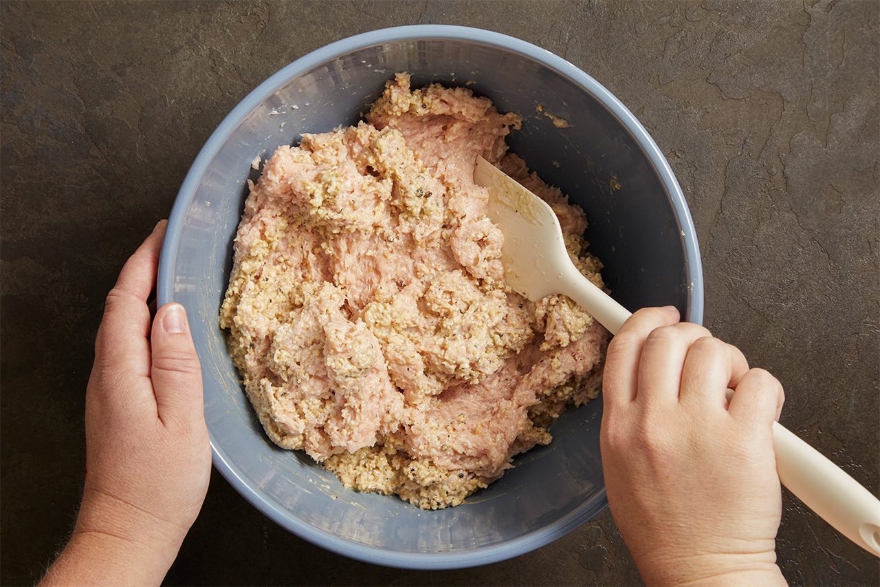Hands mixing raw ground meat and breadcrumbs in a large blue bowl with a white spatula on a dark countertop.
