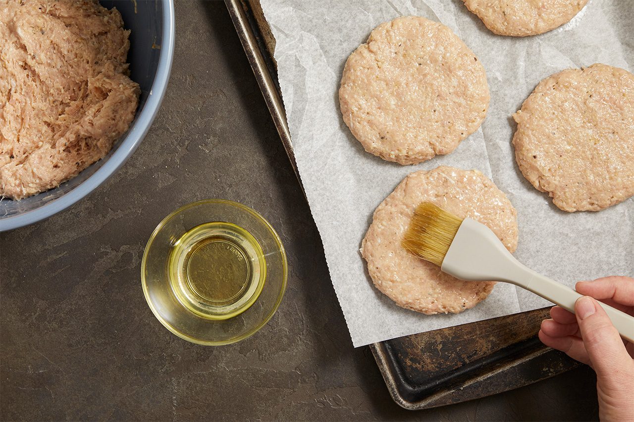 A hand uses a brush to apply oil to round, uncooked patties on a baking tray lined with parchment paper. A bowl of mixture and a small glass bowl of oil are nearby.