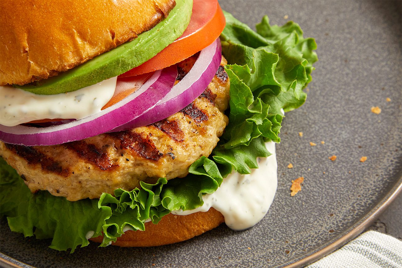 A close-up of a chicken burger with lettuce, tomato, red onion, avocado, and sauce in a bun, served on a gray plate.