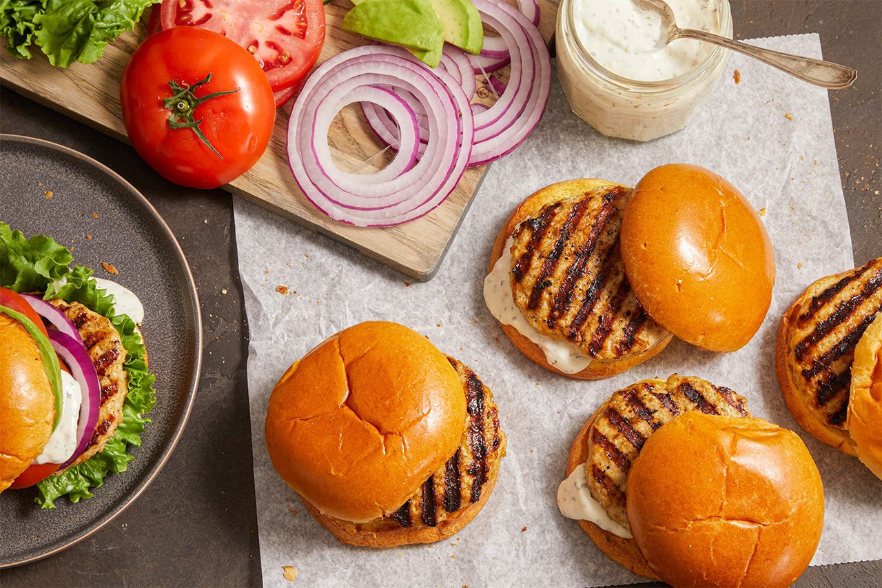 Three grilled chicken sandwiches on buns, with a plate of a sandwich, and sliced tomato, red onion, avocado, and a jar of sauce on a cutting board nearby. Ingredients are arranged on a dark surface.