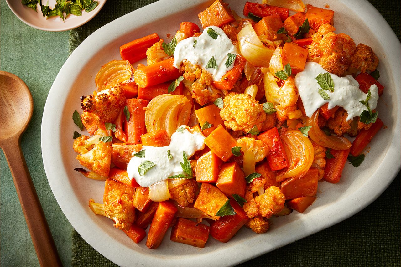 Overhead horizontal image of a white platter filled with roasted carrots, cauliflower, sweet potatoes, and onions. The vibrant vegetables are topped with dollops of a creamy white sauce and garnished with fresh mint leaves. | [Original] RMS 282728 Harissa Roasted Vegetables; Horizontal AP; Pull
