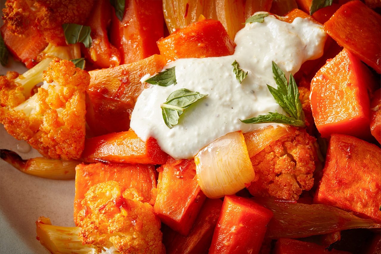 Eye Level horizontal image of a close-up of roasted vegetables, including orange cauliflower florets, cubed carrots, and sliced onions. The dish is topped with a dollop of creamy white sauce and garnished with fresh mint leaves. | [Original] RMS 282728 Harissa Roasted Vegetables; Horizontal; Push