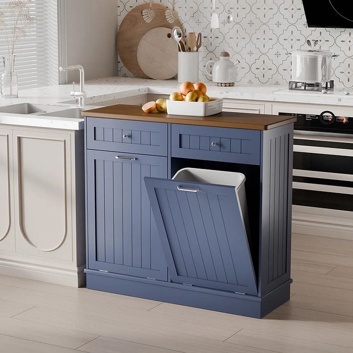 A blue kitchen cabinet with two drawers and a tilt-out trash bin beneath a wooden countertop, holding a white tray of fruit, sits in a modern kitchen setting.