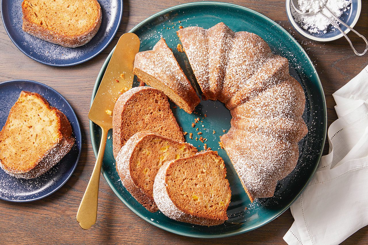 Overhead horizontal image of a bundt cake, partially sliced and dusted with powdered sugar, displayed on a teal serving plate. Two individual slices, also dusted with powdered sugar, rest on smaller blue plates, with a gold cake server beside the main cake. A small bowl of powdered sugar and a sifter are also visible.