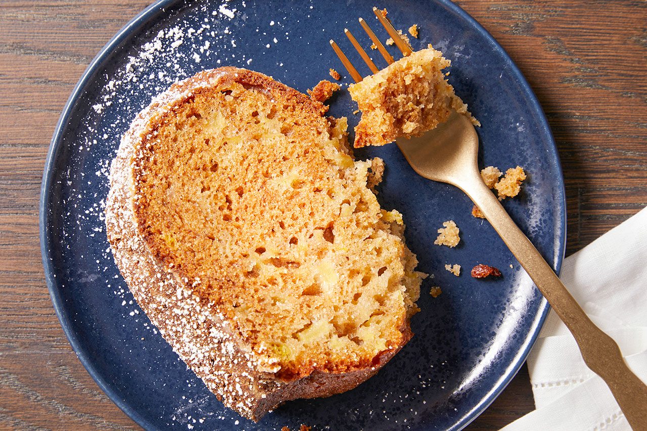 Overhead horizontal image of a slice of cake on a dark blue plate. A golden fork is taking a bite from the cake, which appears to contain pieces of fruit and is dusted with powdered sugar. The plate rests on a dark wooden surface, with a white napkin visible.