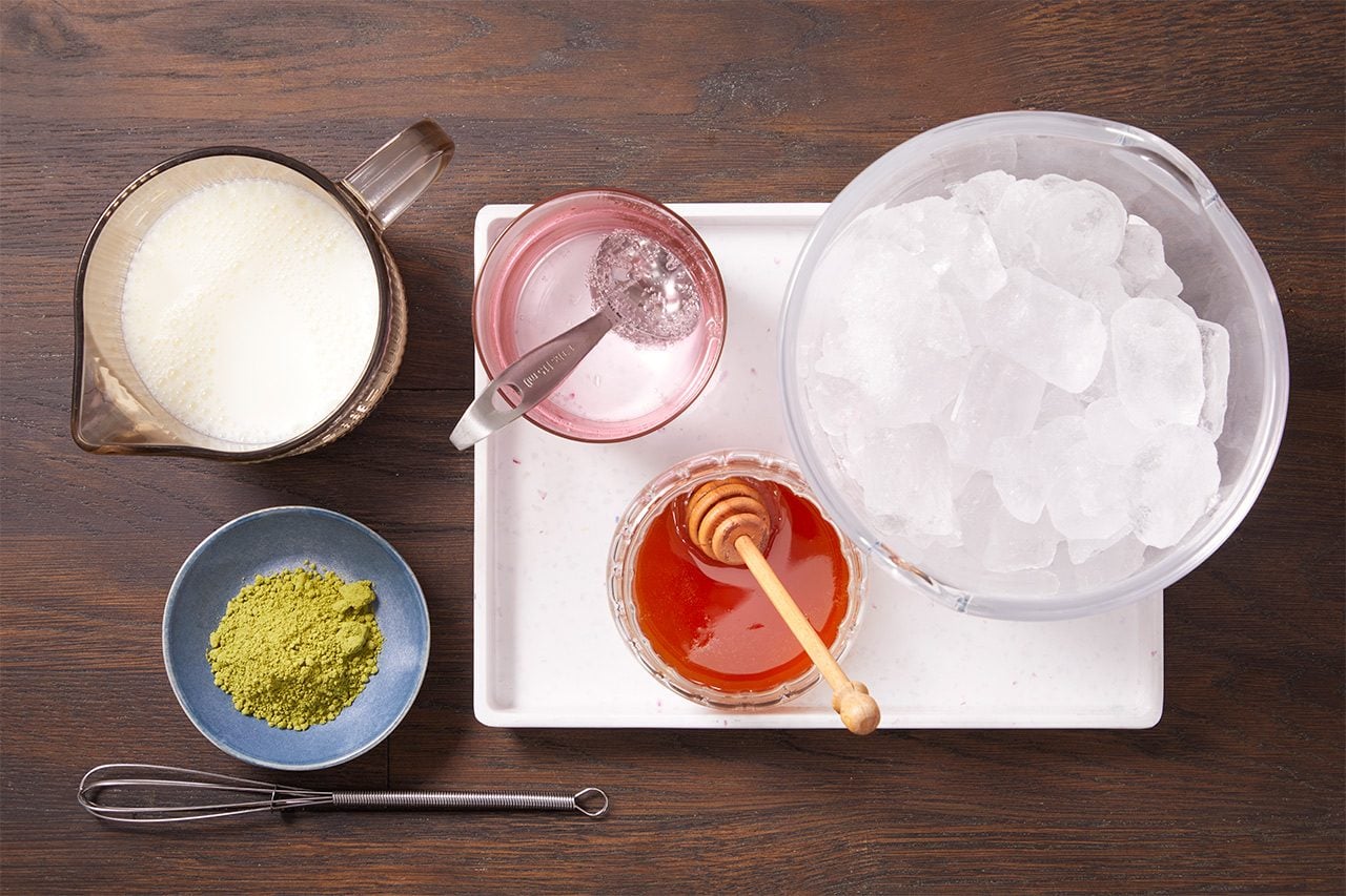 Overhead shot of ingredients arranged for drink preparation, featuring a pitcher of milk, a large bowl of ice, matcha powder, honey with a honey dipper, and a small bowl of pink mixture with a spoon, arranged on a tray over a dark wooden surface.
