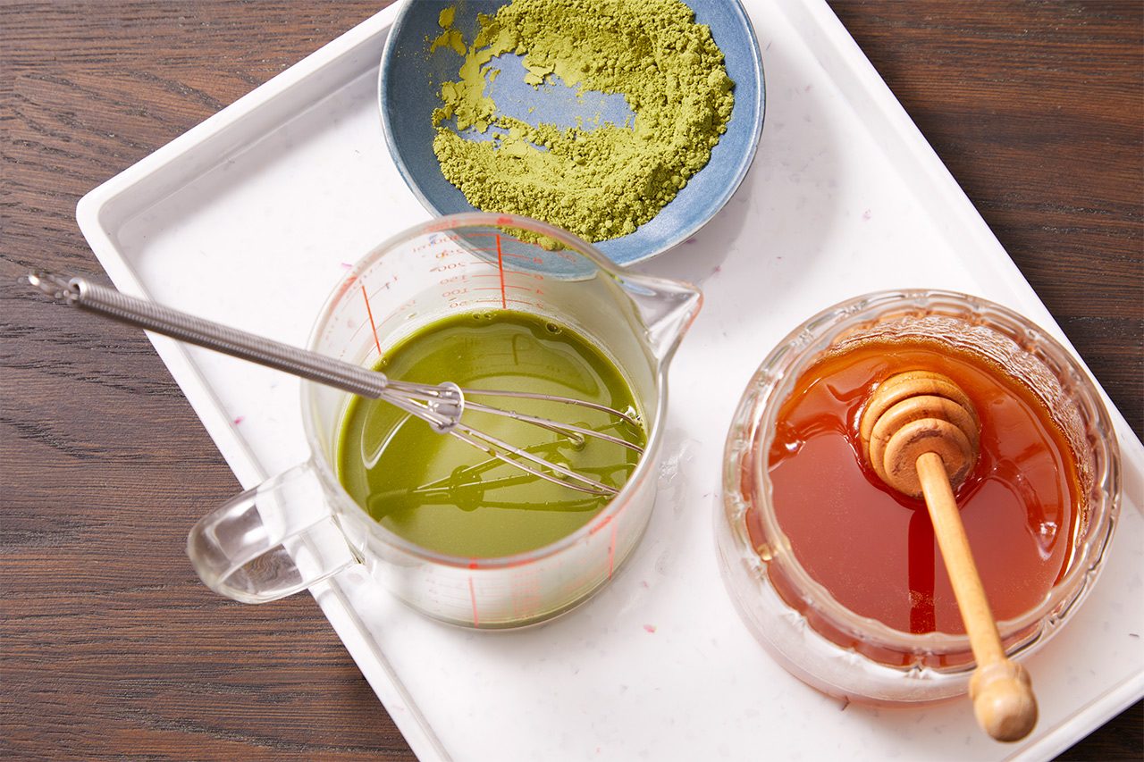Overhead shot of matcha powder being whisked with liquid in a measuring cup, with a bowl of additional matcha powder and a jar of honey with a honey dipper arranged nearby on a white tray over a dark wooden surface.