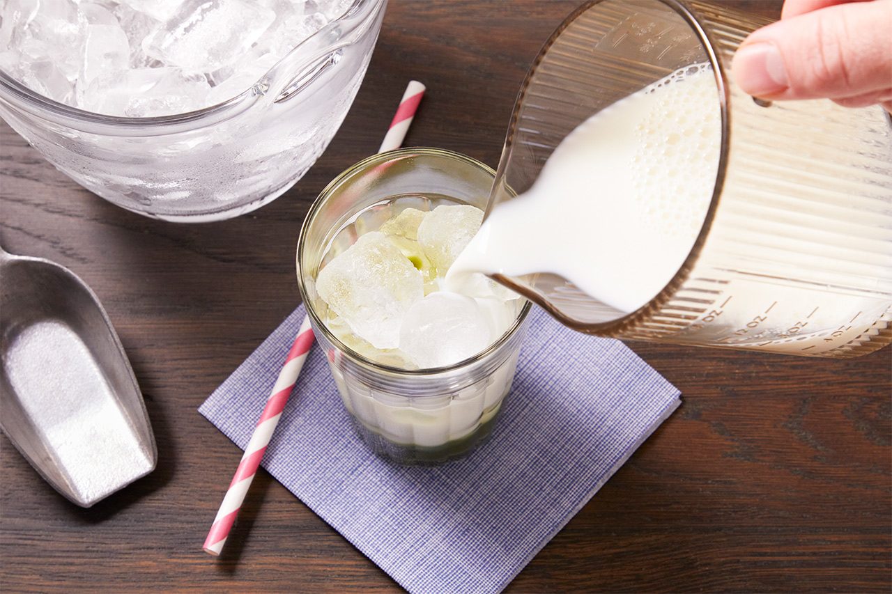3/4 angle view shot of milk being poured from a measuring cup into a glass filled with ice and a green matcha mixture, with a bowl of ice, an ice scoop, and a striped paper straw arranged nearby on a dark wooden surface.