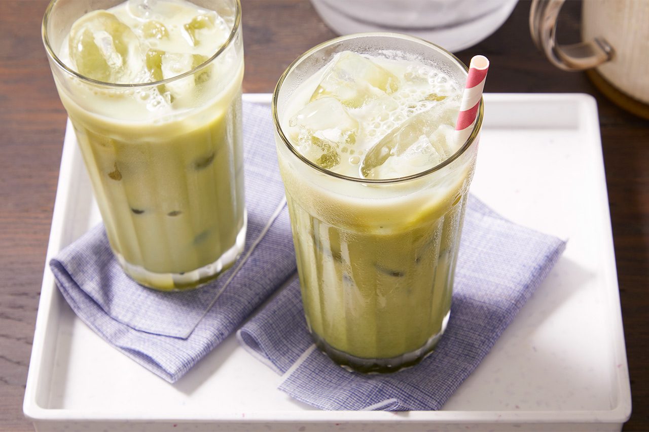 3/4 angle view shot of two Iced Matcha Lattes served in tall glasses filled with ice, with one glass featuring a red-and-white striped straw. The drinks are arranged on blue napkins over a white tray on a dark wooden surface.