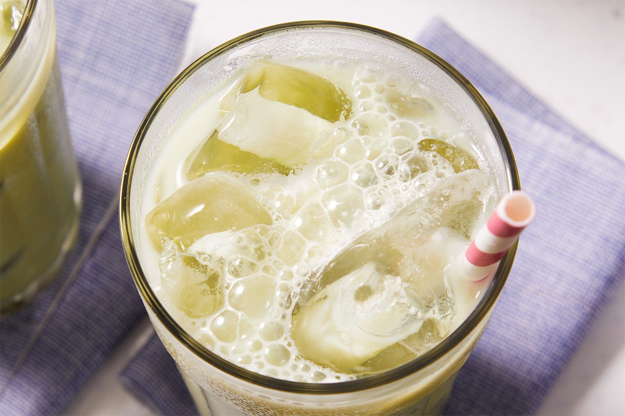 Close-up shot of an Iced Matcha Latte in a tall glass filled with ice, highlighting the creamy, frothy surface and pale green matcha color, with a red-and-white striped straw and a second latte partially visible in the background.