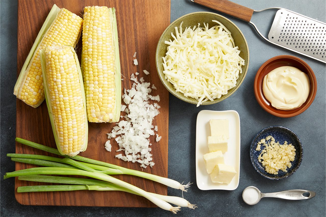Overhead horizontal image of various ingredients laid out on a dark gray surface and a wooden cutting board. The image features three ears of corn on the cob, chopped white onion, green onions, a bowl of shredded cheese, mayonnaise, cubed butter, minced garlic, and a teaspoon of salt.