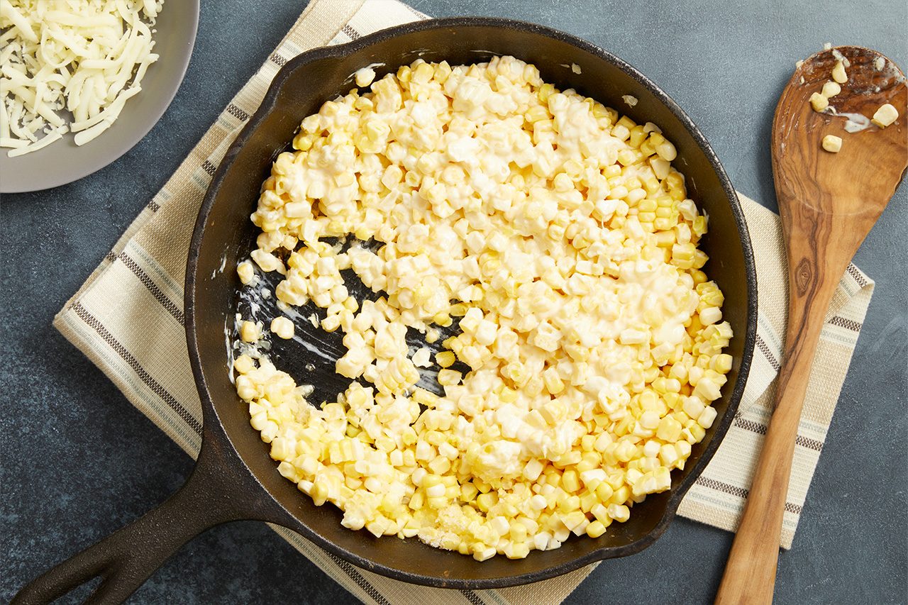 Overhead horizontal image of creamy corn in a black cast iron skillet, positioned on a dark grey surface. A light-colored striped napkin and a wooden spoon are to the right, while a plate of shredded cheese is visible in the upper left corner. The corn kernels are coated in a rich white sauce.