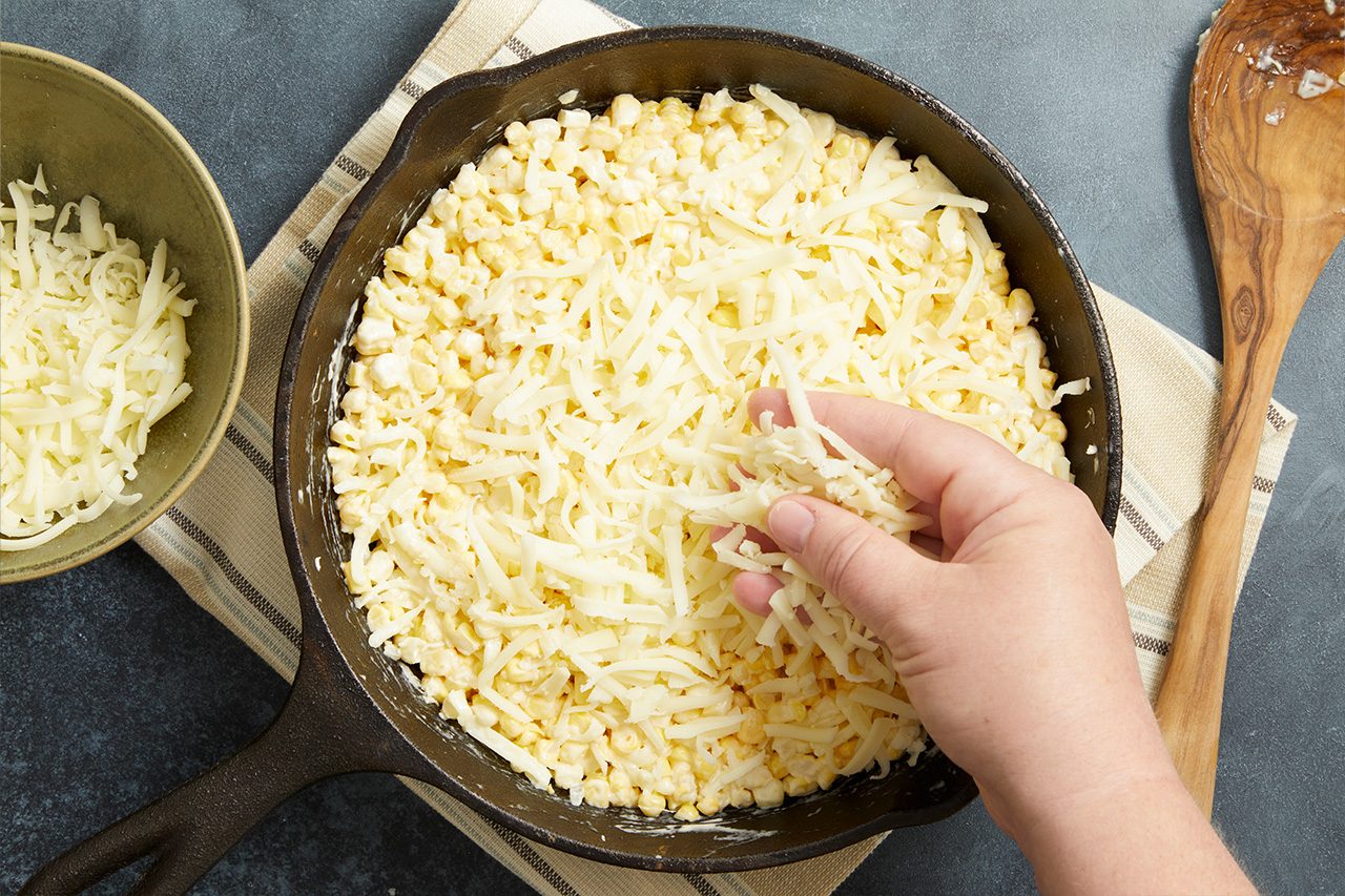 Overhead horizontal image of a hand sprinkling shredded cheese over a mixture of corn kernels in a black cast iron skillet. A bowl of additional shredded cheese sits to the left, and a wooden spoon is visible on a striped napkin to the right.