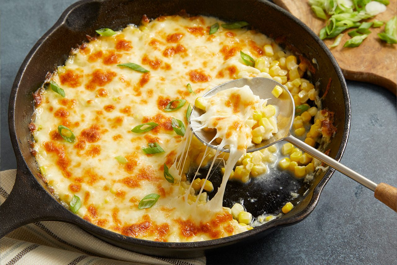 Eye Level horizontal image of a serving spoon scooping cheesy corn from a cast iron skillet. The baked corn dish is topped with bubbly, browned melted cheese and garnished with fresh green onions. More sliced green onions are visible on a wooden cutting board in the background.
