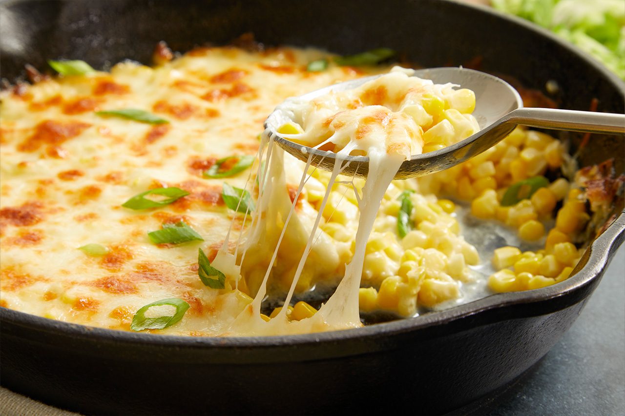 Eye Level horizontal image of a spoon lifting a serving of creamy, cheesy corn from a black cast iron skillet. The melted cheese creates long, tempting pulls as it's lifted, and the dish is garnished with sliced green onions.