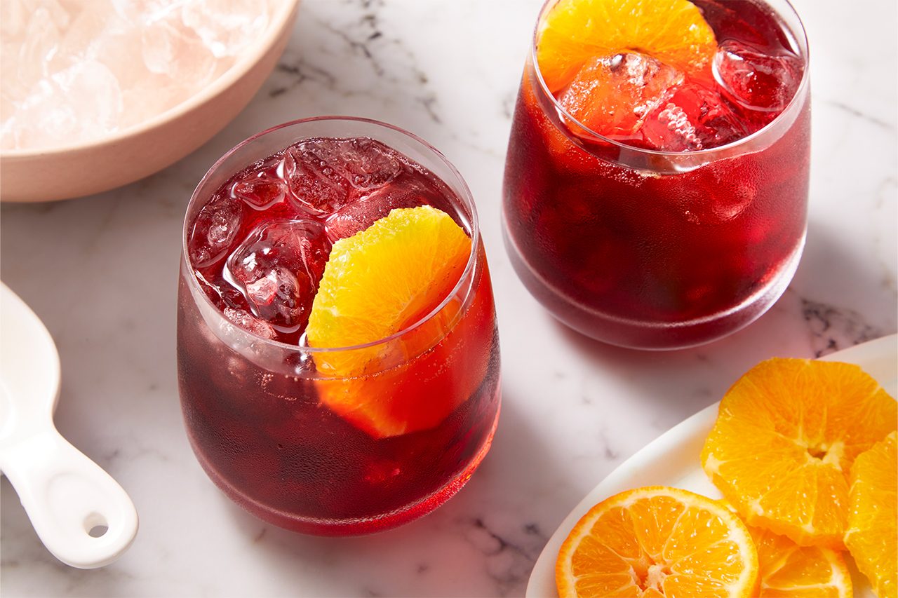 Eye Level horizontal image of two glasses of a dark red beverage, such as sangria, filled with ice and garnished with a fresh orange slice. In the background, a bowl of ice and a plate with additional orange slices are visible on a marble surface.