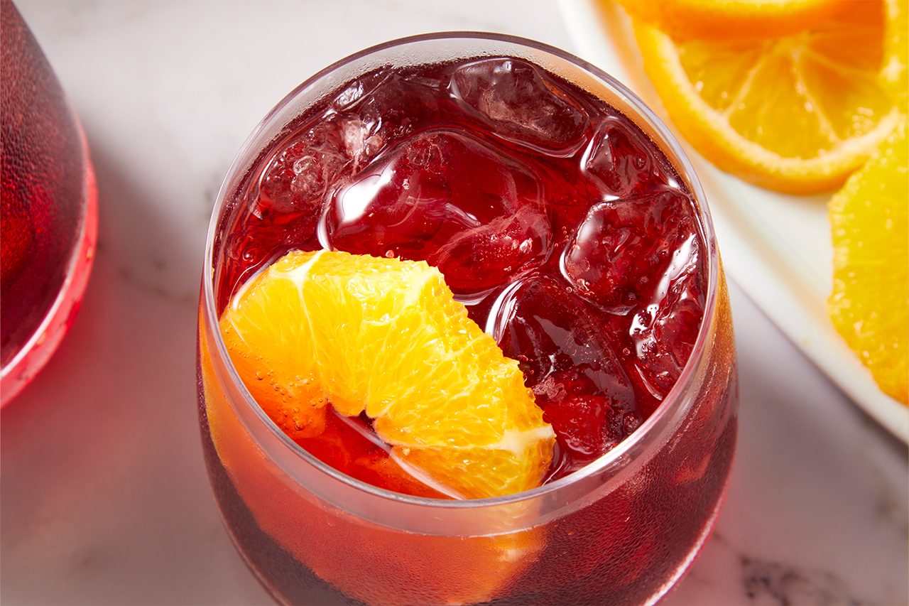 Eye Level horizontal image of a glass filled with a red beverage, ice, and an orange slice. More orange slices are visible in the soft-focus background.