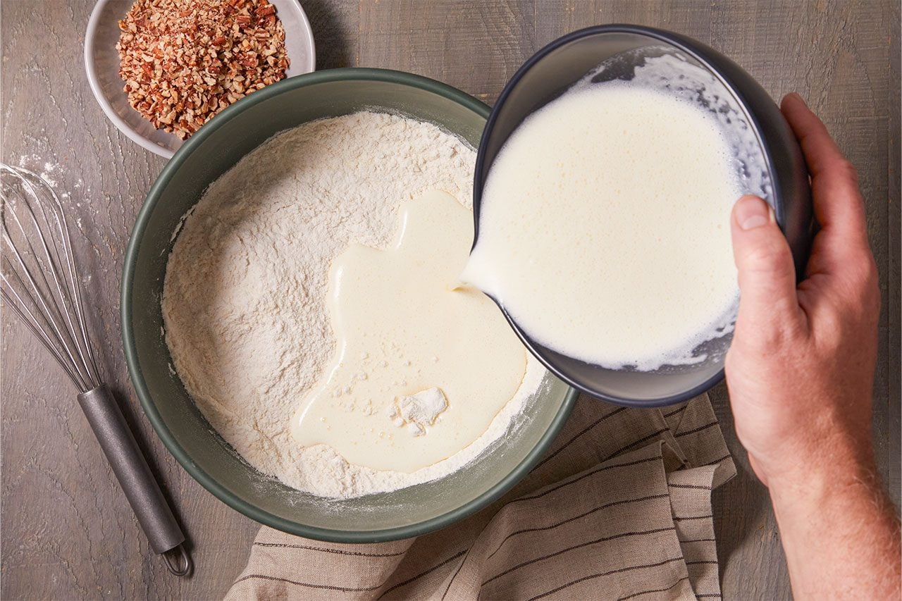 Overhead shot of milk being poured from a bowl into a large mixing bowl of flour to prepare dough, with a whisk, a small bowl of chopped nuts, and a folded kitchen towel arranged nearby on a wooden surface;