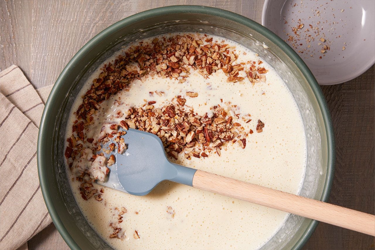 Overhead shot of a large mixing bowl containing a creamy batter as chopped nuts are added on top. A silicone spatula rests in the mixture, with a folded kitchen towel and an empty plate arranged nearby on a wooden surface;