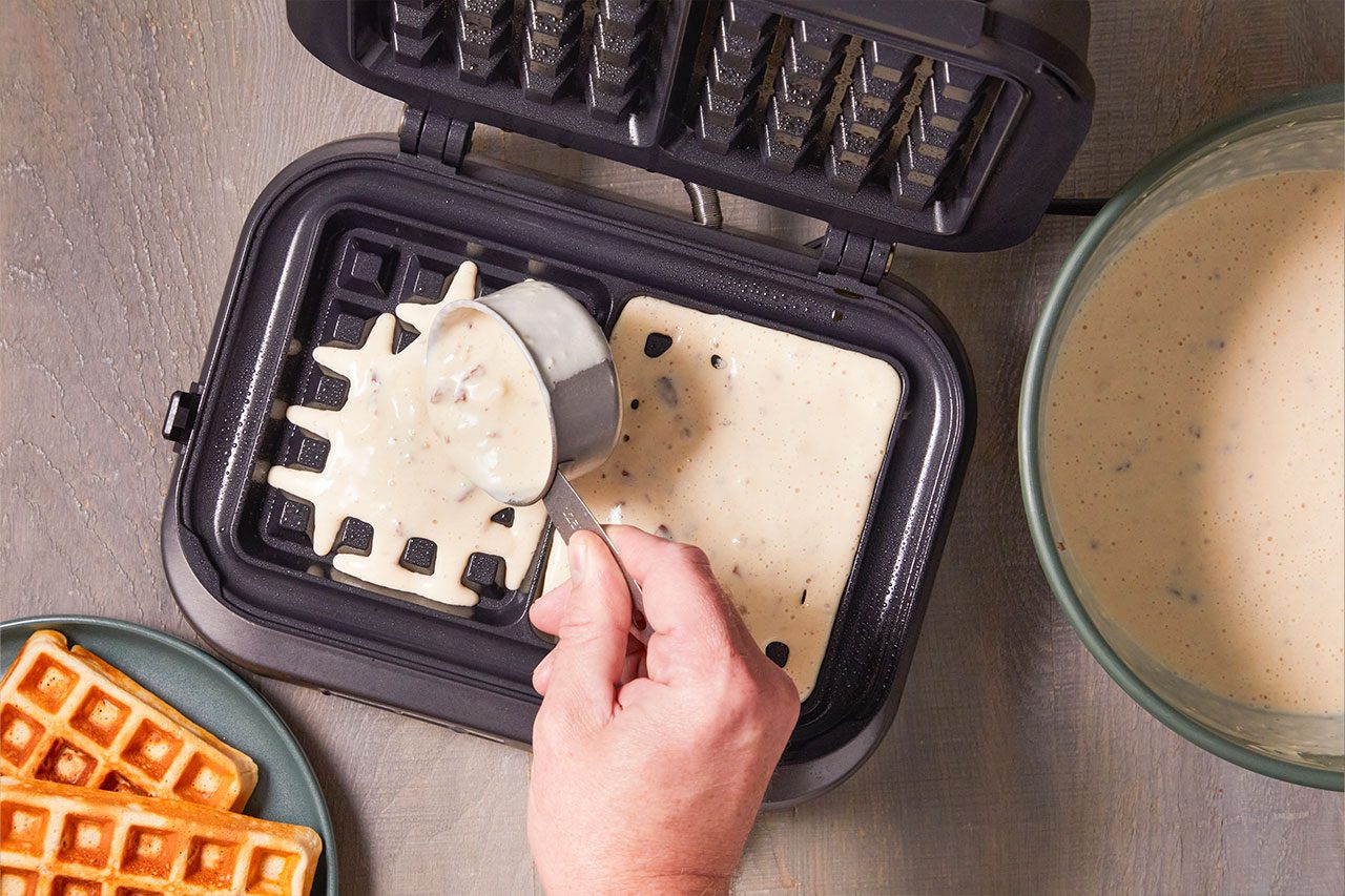 Overhead shot of batter being scooped into an open waffle maker with a measuring cup, while a bowl of prepared batter and a cooked waffle are positioned nearby on a wooden surface;