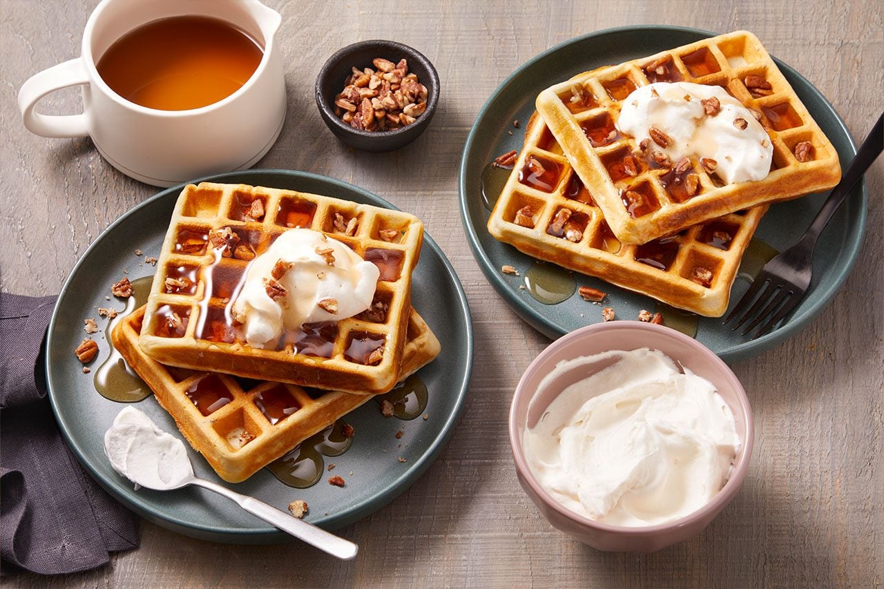 3/4 angle view shot of Malted Butter Pecan Waffles served on two blue plates, topped with whipped cream, chopped pecans, and maple syrup. A mug of maple syrup, a bowl of whipped cream, a small bowl of chopped pecans, and serving utensils are arranged nearby on a wooden surface;