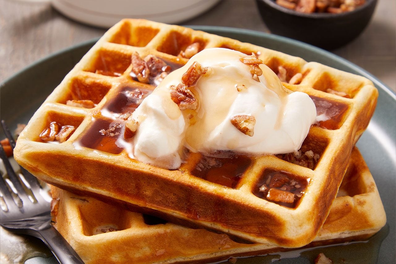 Close-up shot of Malted Butter Pecan Waffles stacked on a blue plate, topped with whipped cream, chopped pecans, and maple syrup, highlighting the crisp golden texture and deep waffle pockets;