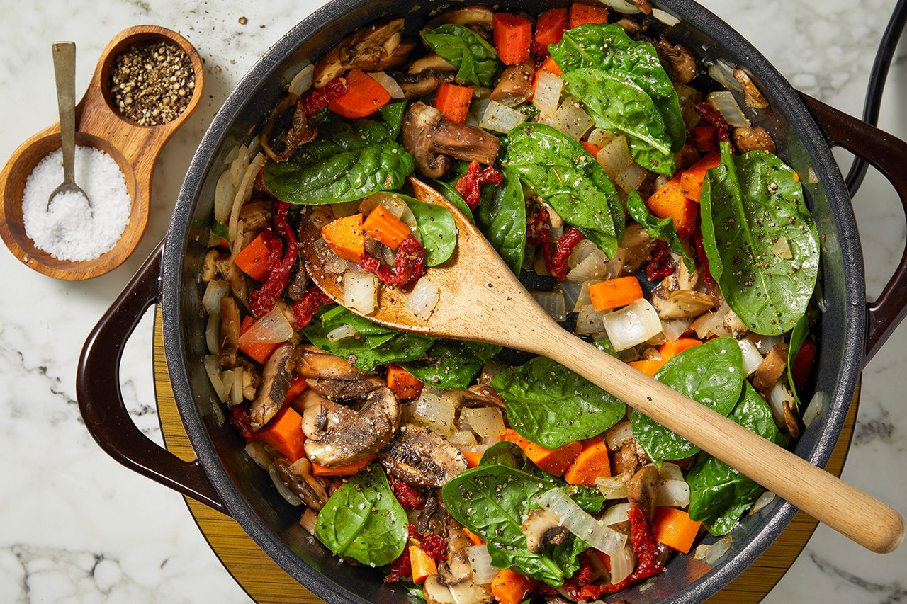 Overhead horizontal image of various vegetables cooking in a dark skillet on a marble surface. Fresh spinach, chopped onions, carrots, mushrooms, and sun-dried tomatoes are visible, mixed with a wooden spoon. A small wooden salt and pepper grinder set sits next to the skillet.