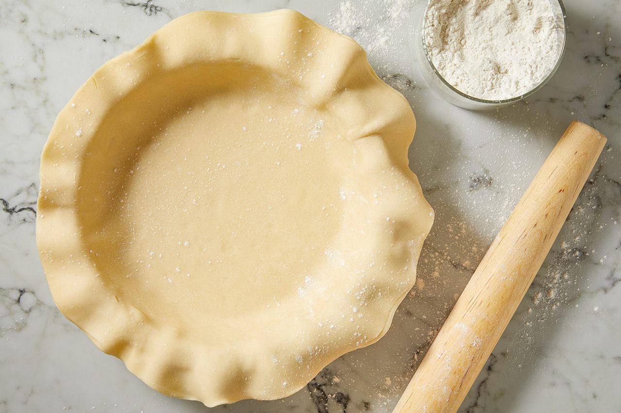 Overhead horizontal image of a raw pie crust pressed into a pie dish, lightly dusted with flour. A wooden rolling pin and a bowl filled with flour are also visible on the white marble surface.