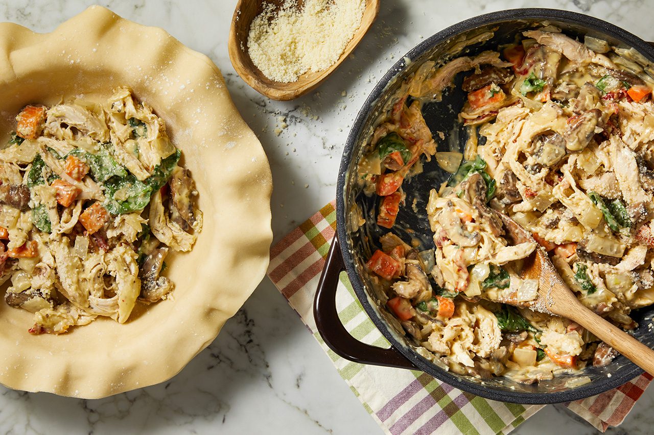 Overhead horizontal image of a pie crust partially filled with a creamy chicken and vegetable mixture on a marble surface. Next to it, a black pan holds more of the filling, which includes shredded chicken, sliced mushrooms, diced carrots, and spinach, along with a wooden spoon. A small wooden bowl of grated cheese is also visible, with some sprinkled over the filling.