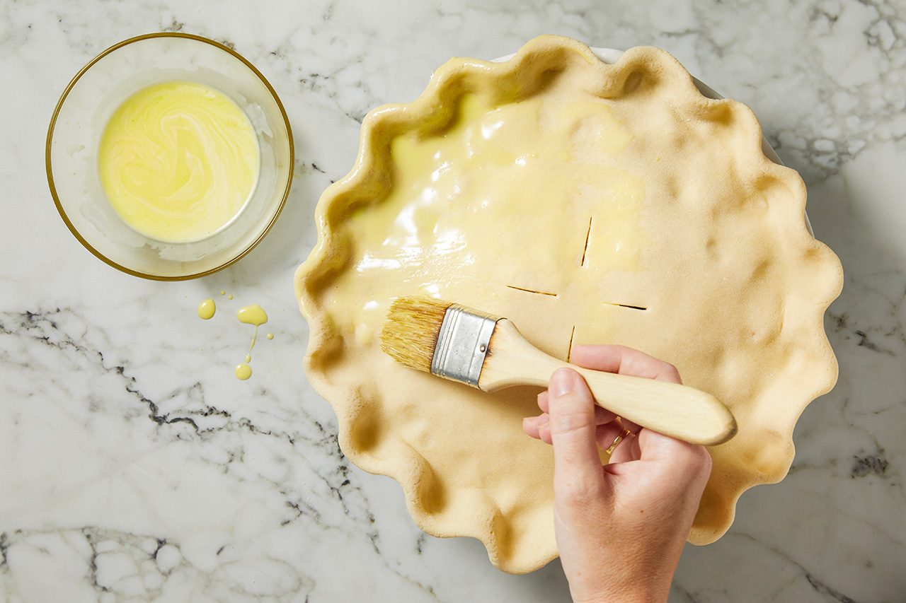 Overhead horizontal image of a hand using a pastry brush to apply a golden liquid, likely an egg wash, onto the top surface of an unbaked pie crust. The crimped pie crust sits in a white pie dish, with a small clear bowl of the liquid positioned to the left.