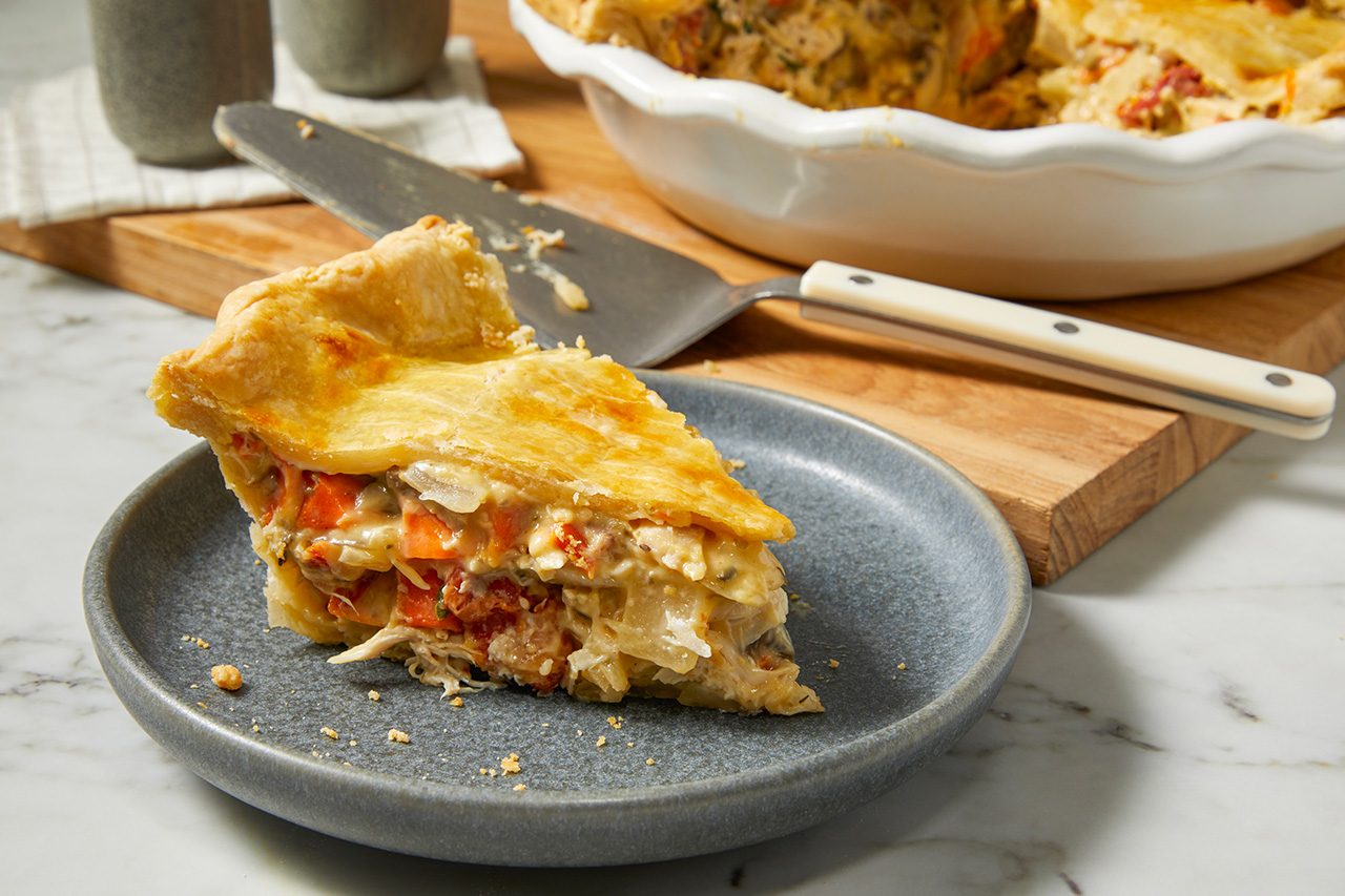 Eye Level horizontal image of a generous slice of savory pie with a flaky golden crust, served on a gray plate. The pie filling features visible pieces of shredded chicken, carrots, and onions in a creamy sauce. In the background, the remainder of the pie sits in a white pie dish next to a pie server on a wooden cutting board.