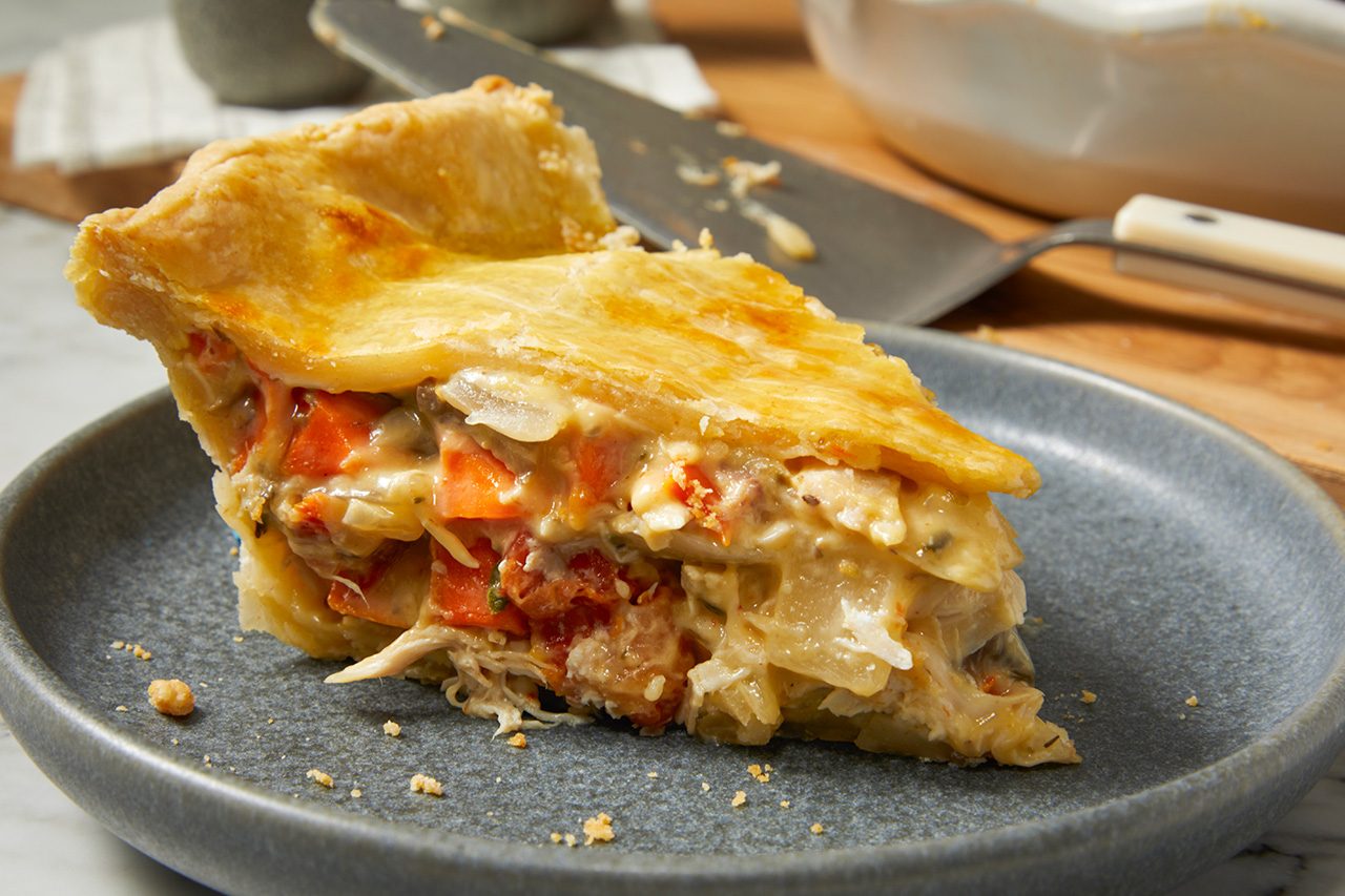 Eye Level horizontal image of a generous slice of savory pie, possibly chicken pot pie, resting on a gray, textured plate. The golden-brown flaky pie crust encases a creamy filling rich with shredded chicken, diced carrots, and sliced onions. In the soft-focus background, a pie server and the edge of a white pie dish are visible.