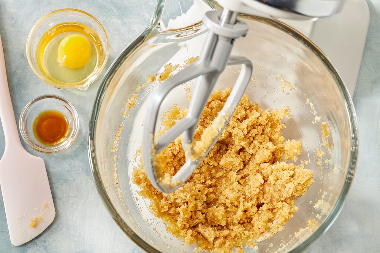 Top-down image of melted butter combined with brown sugar and granulated sugar in a mixing bowl using an electric mixer, beginning the cookie dough base.