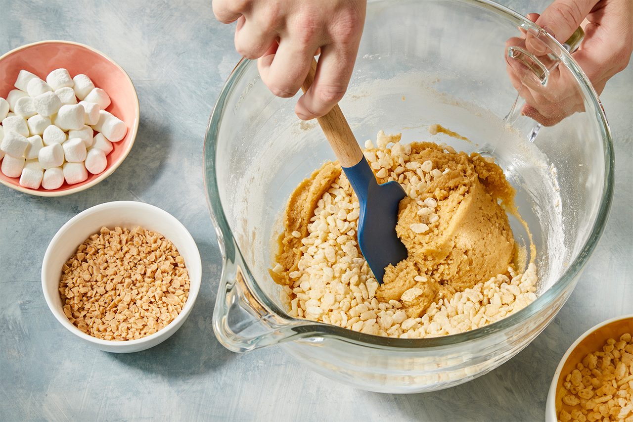 Top-down image of Rice Krispies cereal, miniature marshmallows, and toffee bits being folded into cookie dough using a spatula.