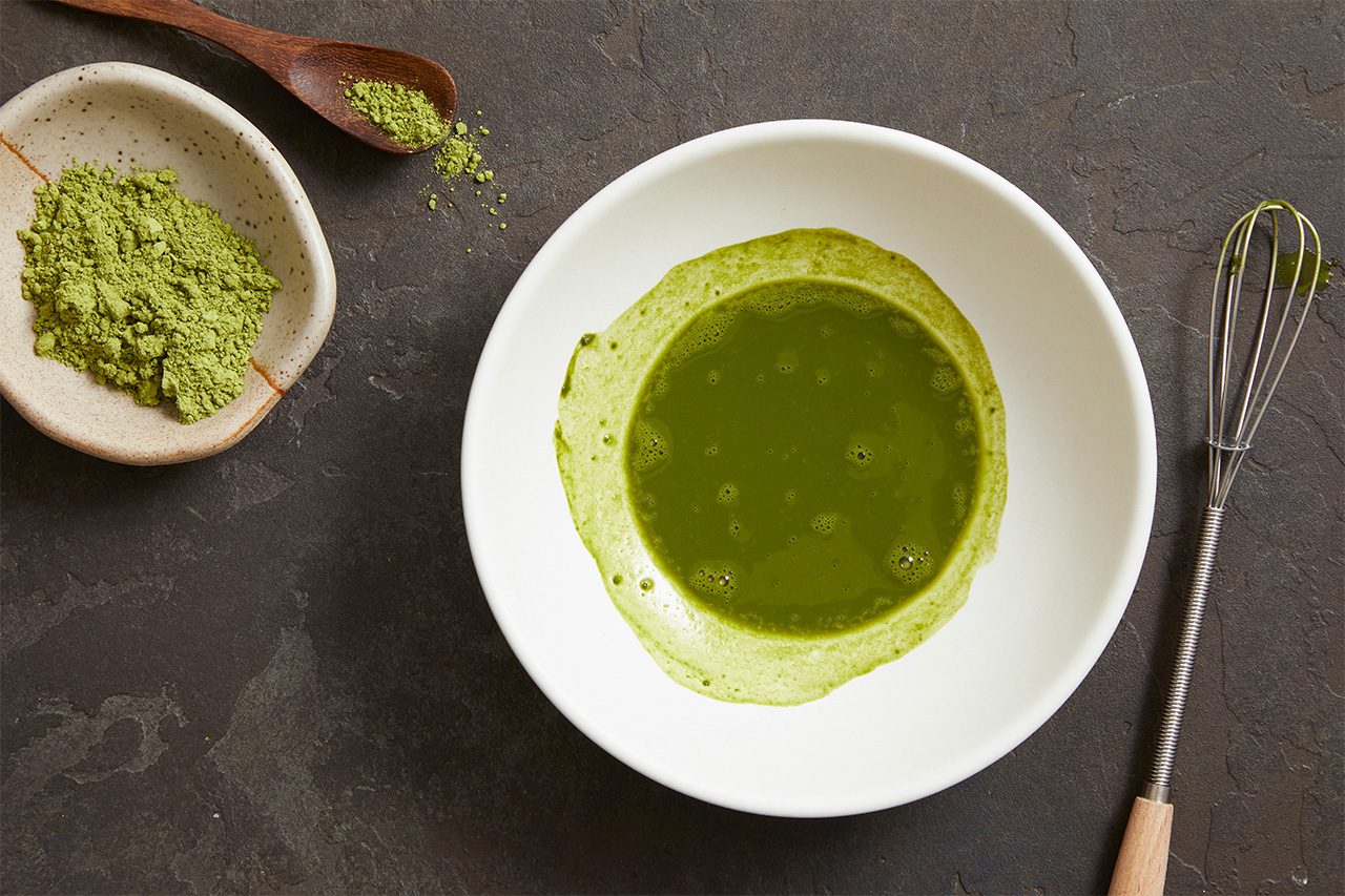 Overhead horizontal image of a white bowl containing whisked green matcha tea with a frothy edge. To the left, a small ceramic bowl holds vibrant green matcha powder, with a wooden spoon holding more powder next to it. A metal whisk with a wooden handle rests to the right of the liquid-filled bowl.