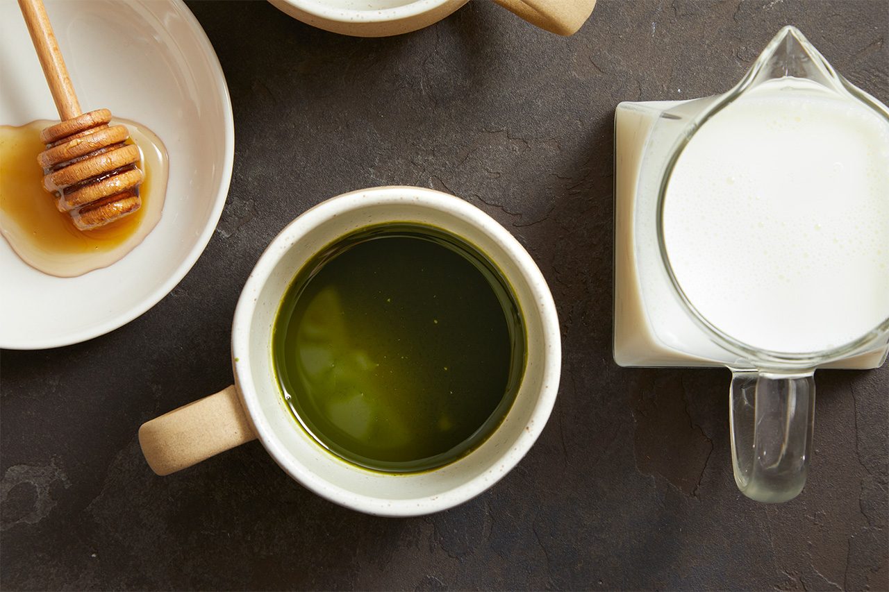 Overhead horizontal image of ingredients laid out on a dark, textured surface. A white mug containing vibrant green matcha tea is centrally placed, with a small white plate holding a honey dipper in honey to its left, and a glass measuring cup filled with milk to its right.