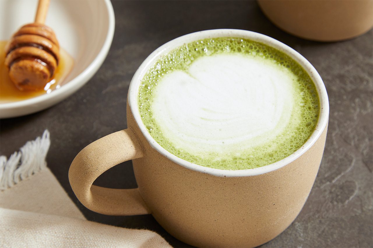 Eye Level horizontal image of a ceramic mug filled with a vibrant green matcha latte, topped with frothy white milk foam in a delicate design. A bowl of honey with a wooden dipper is visible in the background, next to a white linen napkin.