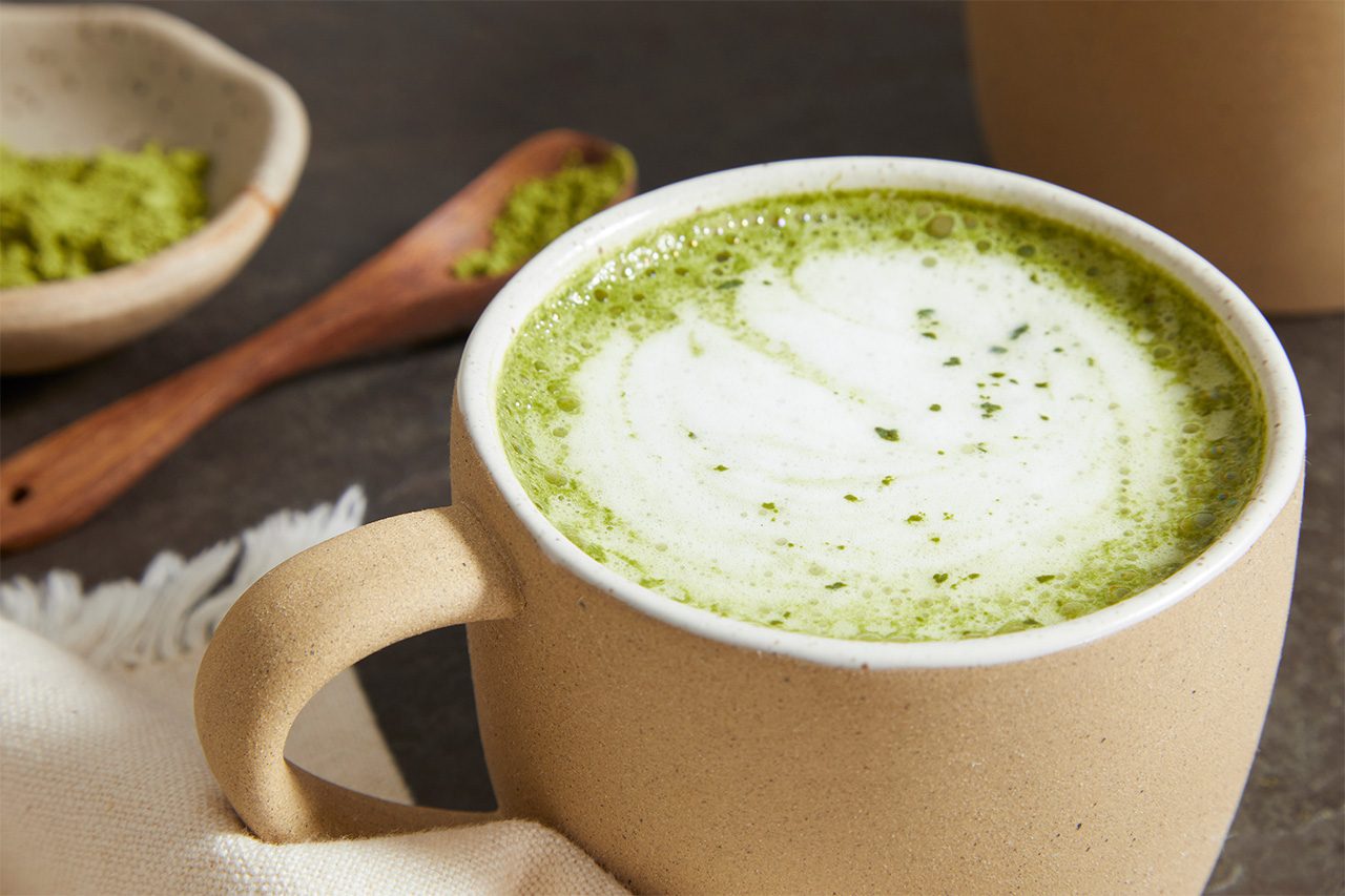 Overhead horizontal image of a creamy matcha latte served in a textured beige mug. The vibrant green matcha drink is topped with a swirl of white foam and a dusting of matcha powder. In the background, a small bowl and a wooden spoon both hold additional matcha powder.