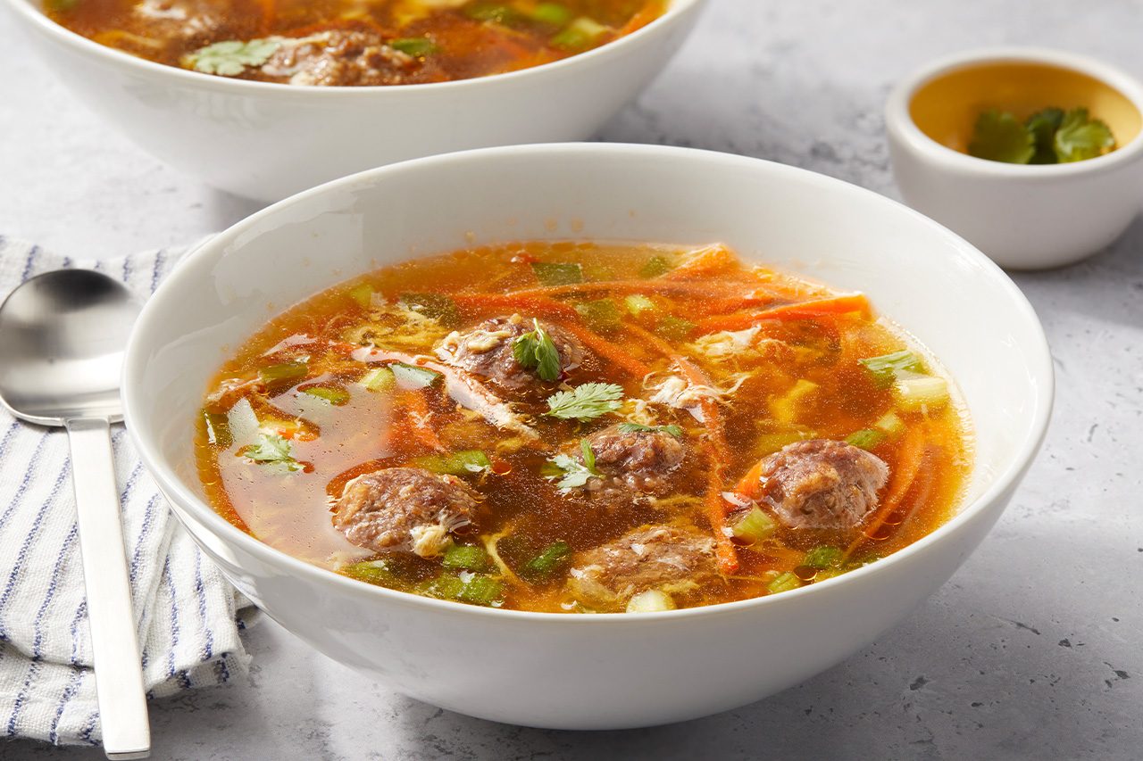 Eye Level horizontal image of two bowls of savory soup, with the front bowl in sharp focus. The clear broth contains meatballs, shredded carrots, chopped green onions, and egg drops, garnished with fresh cilantro. A stainless steel spoon rests on a striped napkin next to the front bowl, while a small dish with additional cilantro is visible in the background. | [Original] RMS 282644; Thai Meatball Egg Drop Soup; Horizontal AP; Pull