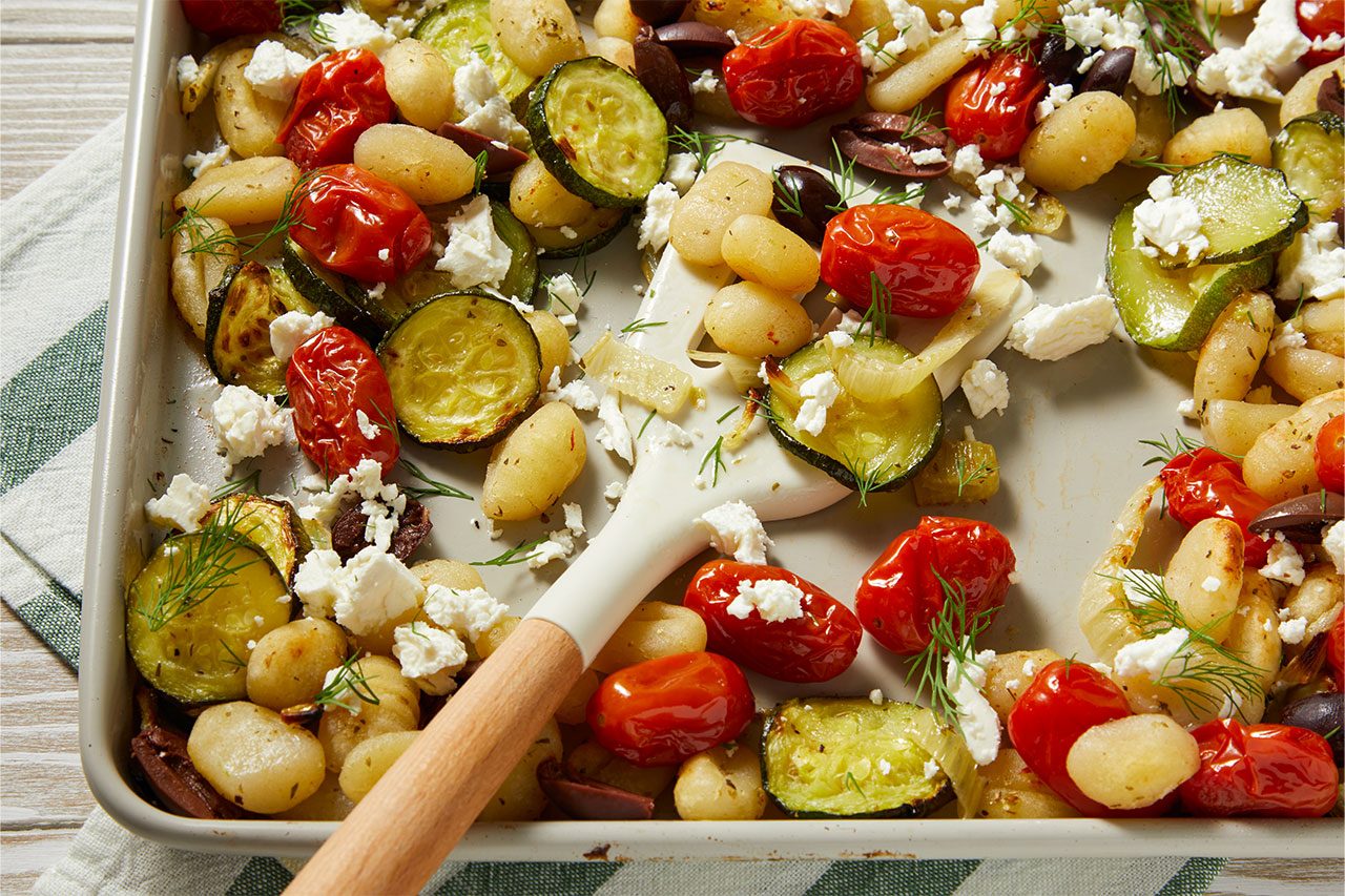 Close-up shot of Mediterranean Sheet-Pan Gnocchi featuring roasted gnocchi with zucchini, cherry tomatoes, red onion, and olives, topped with crumbled feta cheese and fresh dill. A serving spoon rests on the sheet pan, highlighting the roasted vegetables and Mediterranean flavors.