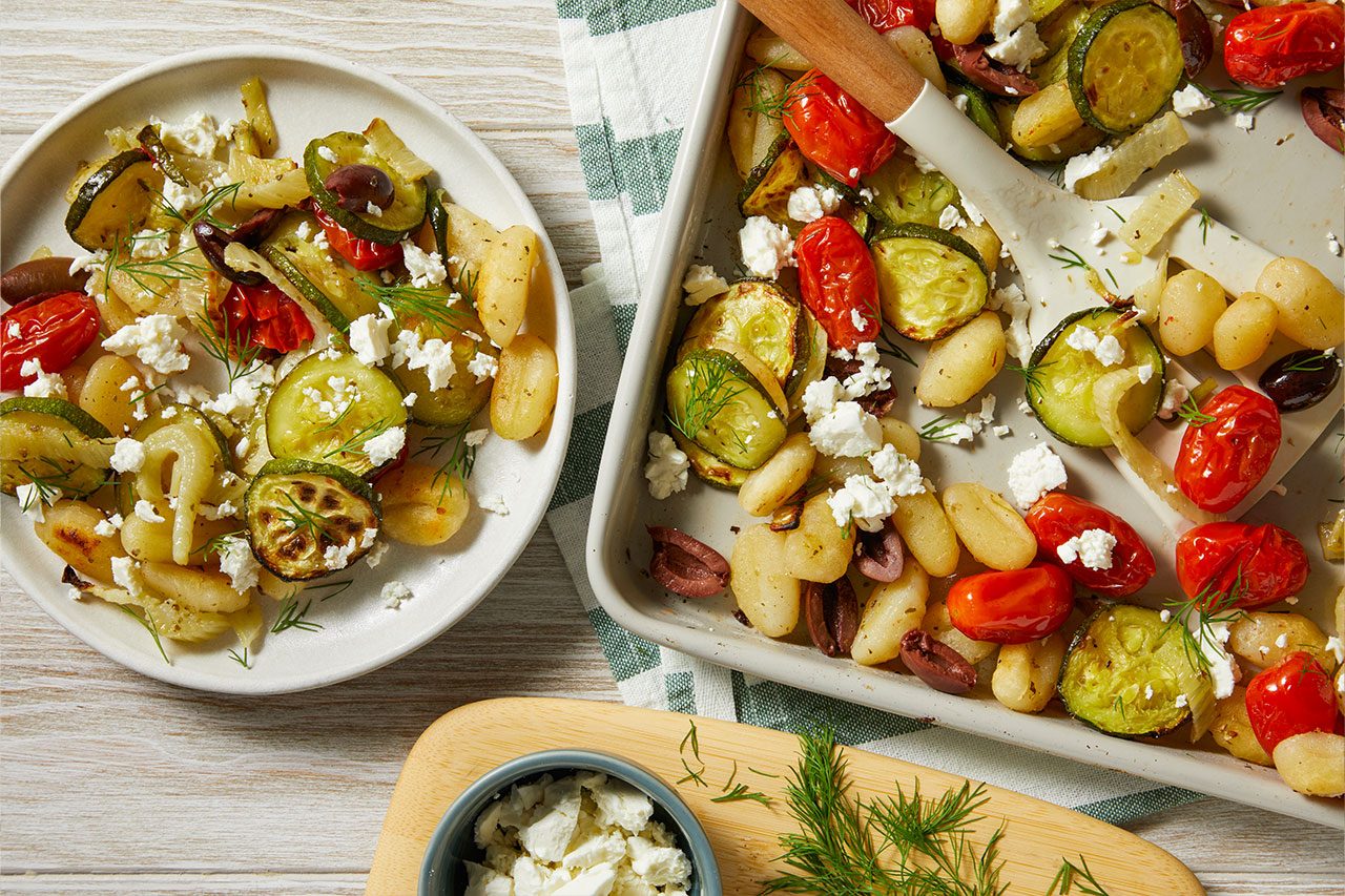Overhead shot of Mediterranean Sheet-Pan Gnocchi served on a white plate alongside the sheet pan, featuring roasted gnocchi with zucchini, cherry tomatoes, red onion, and olives, topped with crumbled feta cheese and fresh dill. A serving spoon rests on the sheet pan, with extra feta and fresh dill arranged nearby.
