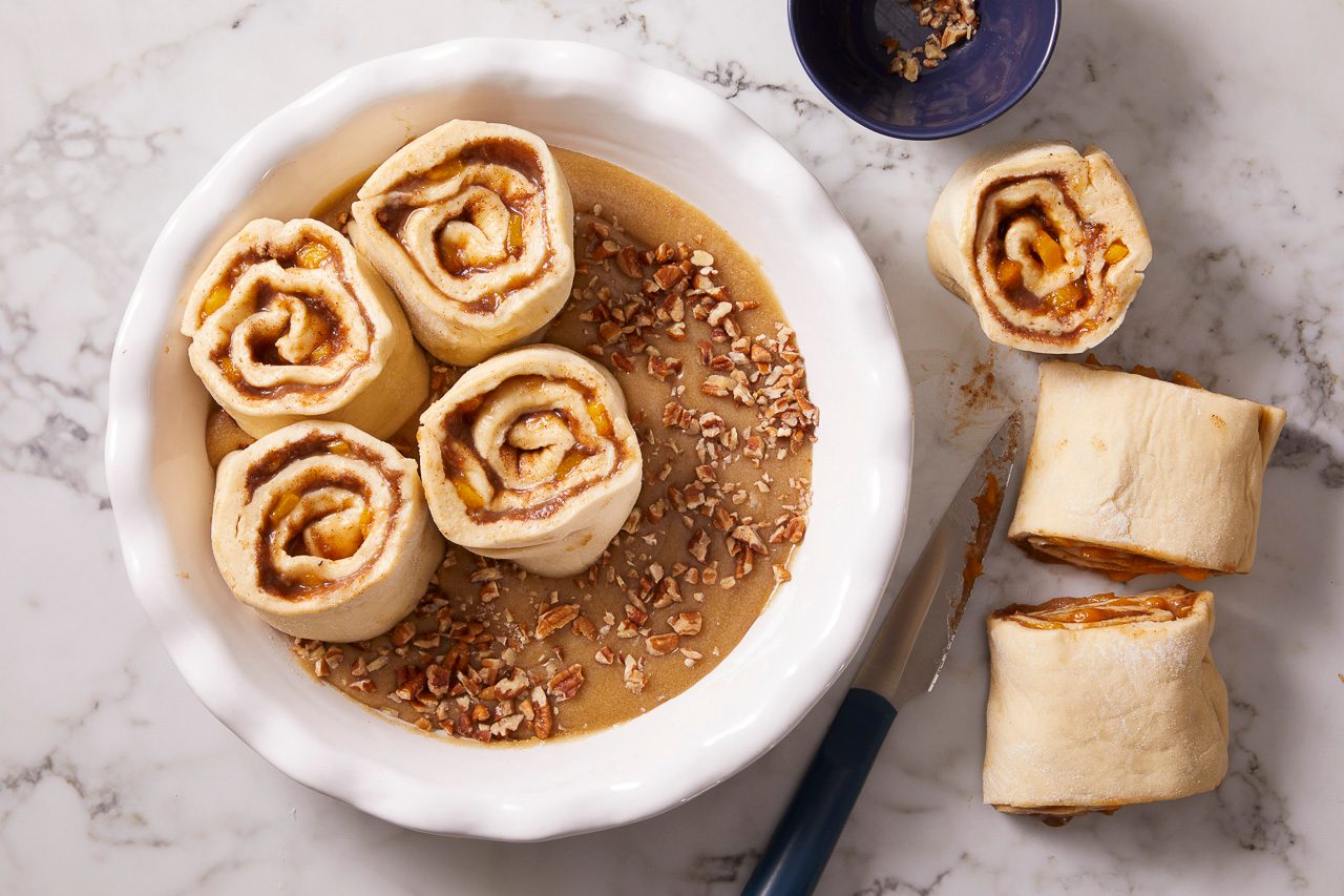 Overhead shot of sliced fruit-filled dough rolls arranged in a buttered baking dish, with chopped pecans sprinkled over the top.
